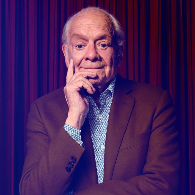 David Jason holds his hand to his chin with a slight smile, dressed in a suit in front of a red curtain.