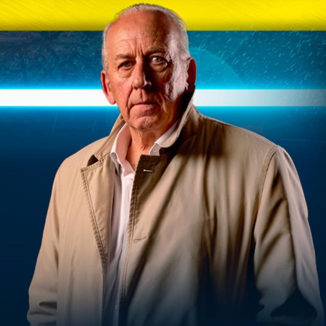 Peter Bleksley stands in a trench coat in front of a bright blue light and rainy brickwork. 