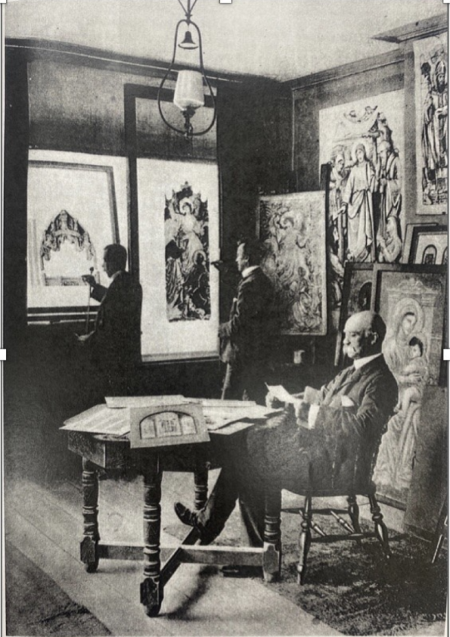 Historic black-and-white photograph of an art studio or gallery. One person sits at a table reviewing papers while two others examine framed religious artworks displayed on the walls, including paintings and icons leaning against the room's wood-panelled interior.