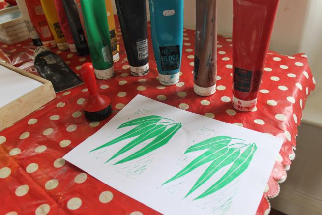 Image shows lino printed leaves with colourful ink tubes in the background on a dotty red tablecloth.