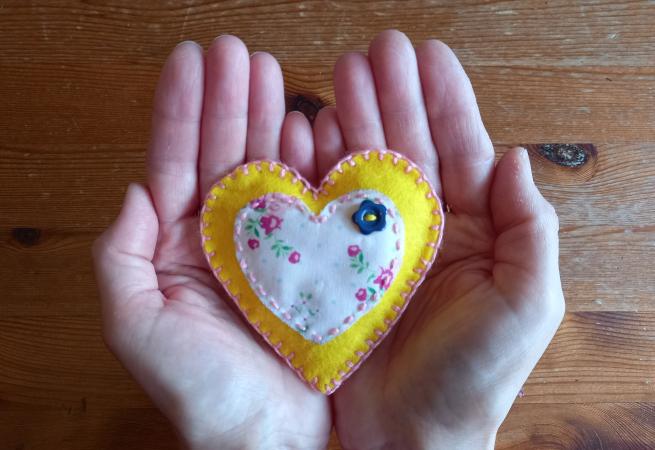 Two hands cupped together holding a fabric heart made from yellow fabric with a smaller flowered fabric heart shape stitched on to it.