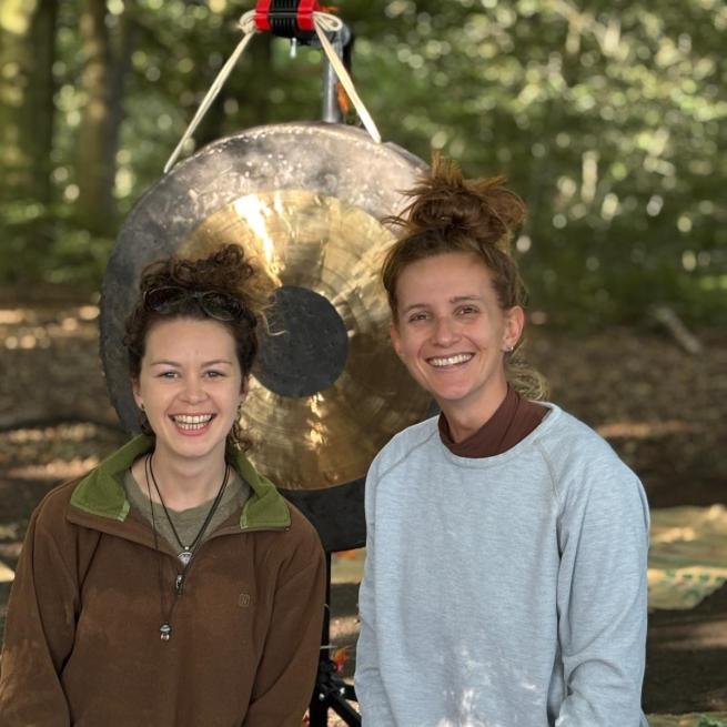 2 women smiling in a woodland in front of a gong