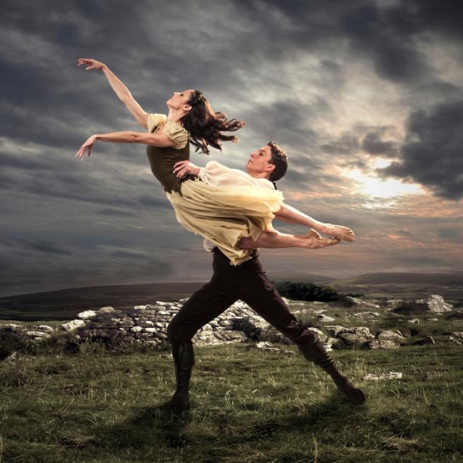 Two ballet dancers in period costume perform a lift out in the moors, with rocks and a cloudy sunrise in the background.