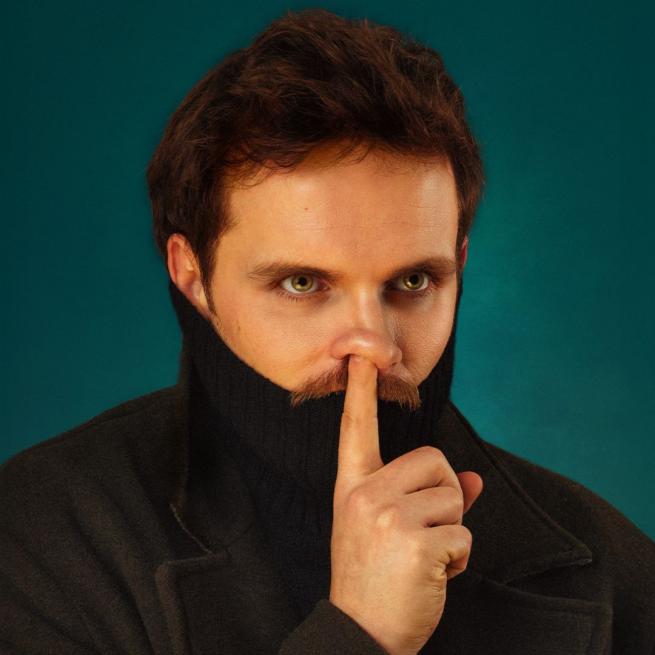 Vittorio Angelone with a turtle neck pulled up over his mouth and his finger over where is mouth would be in a 'be quiet' gesture.