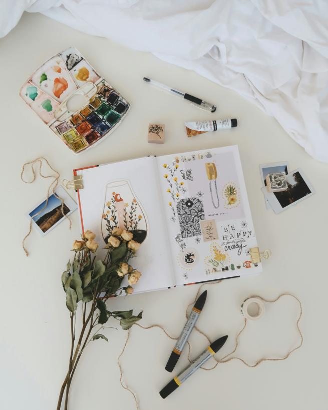 Open creative journal with collage art, dried roses, paint palette, pens, and craft supplies arranged on a white tabletop.