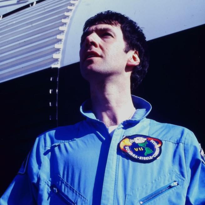 Clint Boon in blue overalls and a patch with a logo of a spaceship. He looks up, off-camera, standing in front of a white and black rocket.