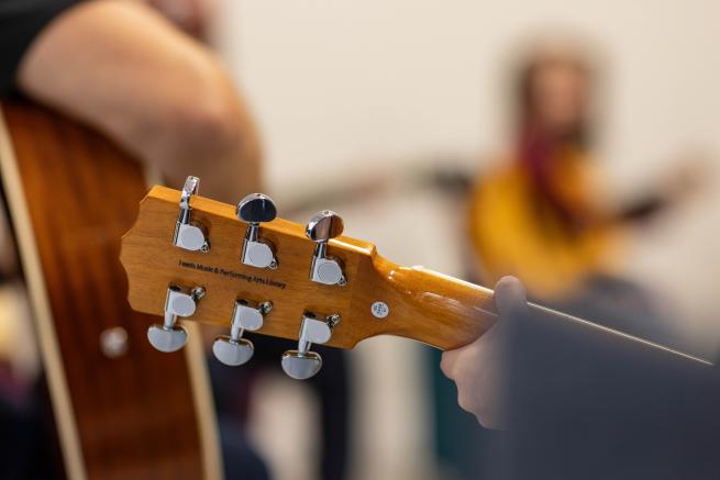 The foreground shows the tuning pegs of a guitar, in the background are blurred figures of guitar players holding guitars.