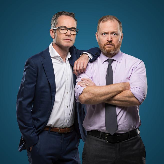 Two men in suits and office attire stand straight-faced against a blue background. One has his arms crossed, the other rests his arm on his shoulder.