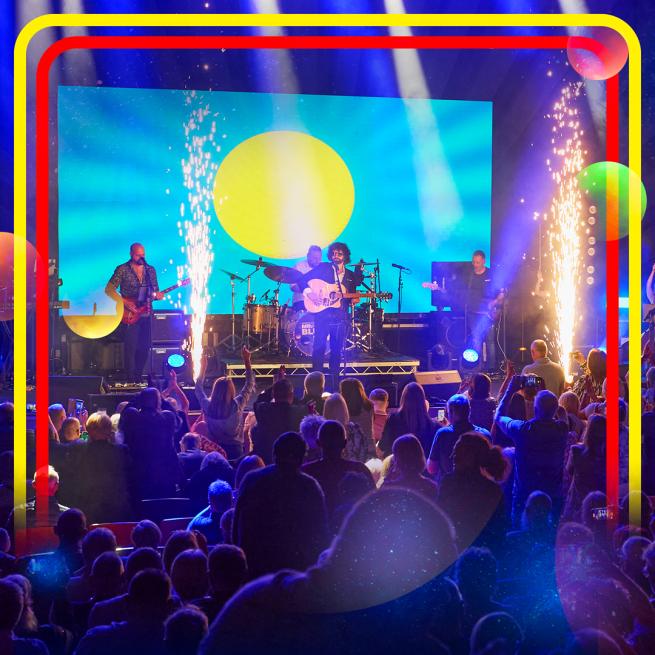 A band performs to a crowd as gold sparks shoot from the stage. A spaceship soars through a night sky.