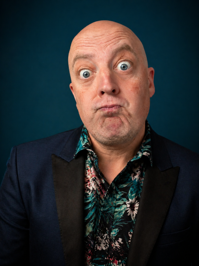Portrait of Antony Vila wearing a dark jacket and patterned shirt, pulling a playful expression against a dark teal background.