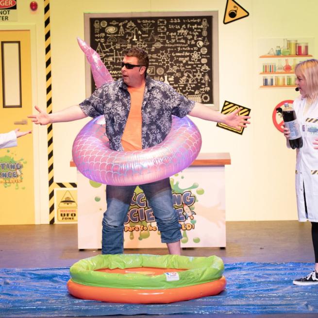 Man wearing an open shirt and a pink swimming rubber ring stood behind a small paddling pool with a blackboard with science-y things written behind him and someone in a lab coat next to him.