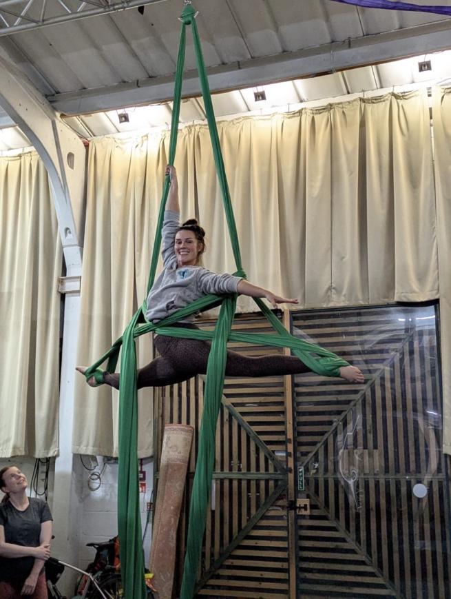 Jess is hanging split legged on the aerial silks at open source arts, a good 10 ft high, but smiling and posing calmly at the camera. Jess is being watched from below by one of her class attendents