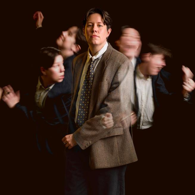 Chloe Petts stands in a tweed jacket, shirt and tie, surrounded by four blurred versions of herself in motion. 