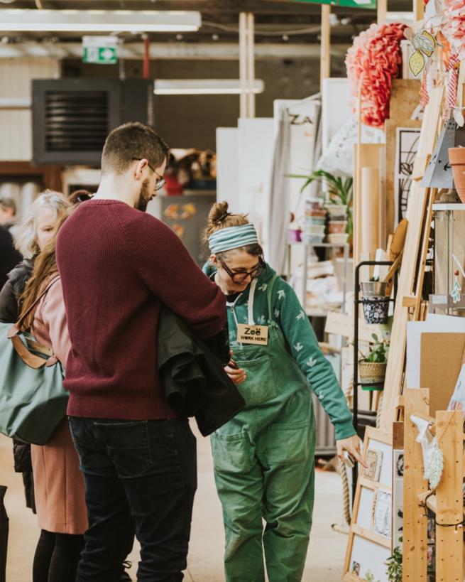 studio holder Zoe wearing green overalls and a headband talking to a pair of visitors in the corridor of her studio space, which is filled with art materials and plants. 