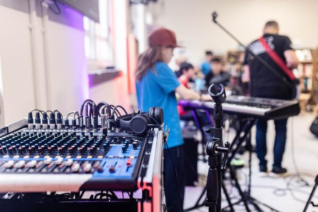 A sound mixing deck sits in the foreground, young people are in the background playing instruments such as a keyboard.