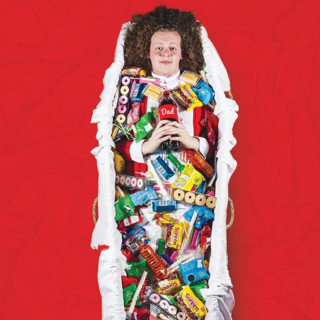 Jack Rooke lies in a coffin-shaped basket full of sweets and biscuits, holding a cola bottle with a label reading 'Dad', surrounded by red material.