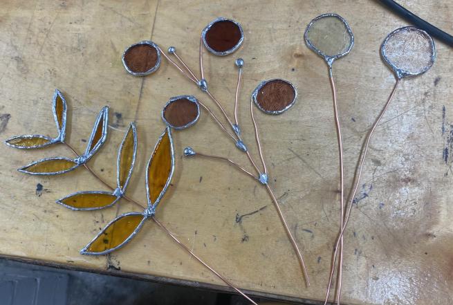 A photograph of three flower stems made of silver wire and stained glass. These are laid upon a wooden workbench