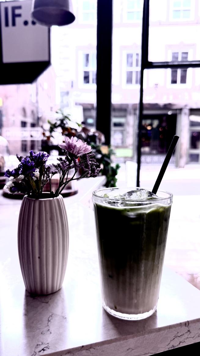 Lilac-tinted vertical shot in close-up of an iced drink in a glass with two straws, next to a small vase with purple cut flowers. Both are in the foreground. In the background there is a hung signage ‘IF’ at the top left corner of the image. 
