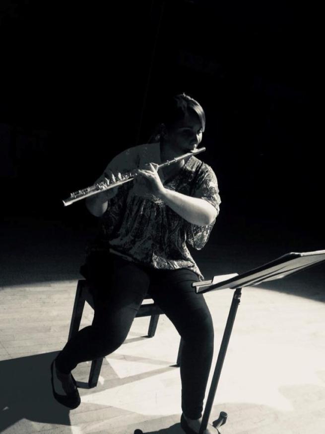 A lady sits on a chair on an otherwise empty stage playing flute portrait of a flautist in black and white