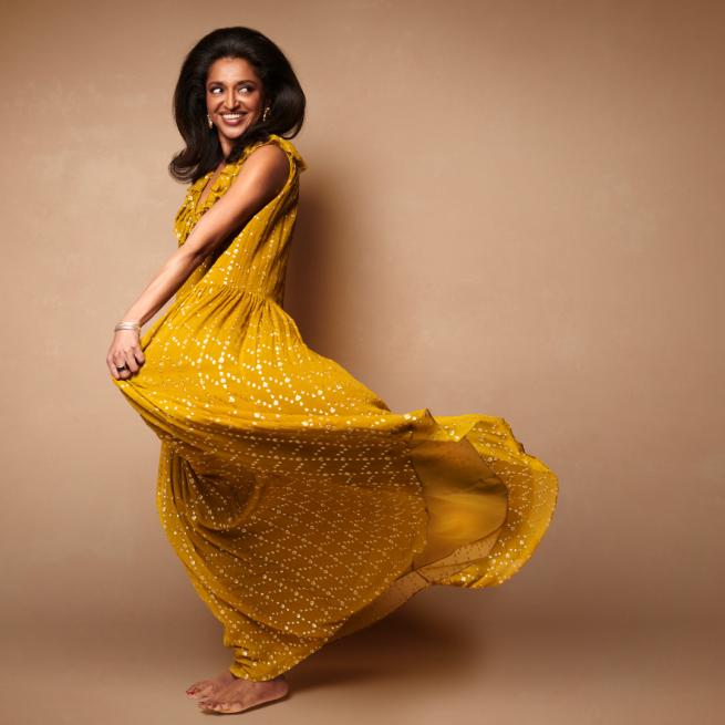 Sindhu Vee spins around a flowing yellow dress and looks behind her shoulder with a smile in front on a pale brown backdrop. She has a blowout hairdo and gold earrings and bangle.