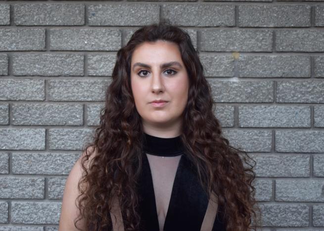 A lady with long wavy dark hair stares enigmatically into the camera, she is stood in front of a plain grey wall