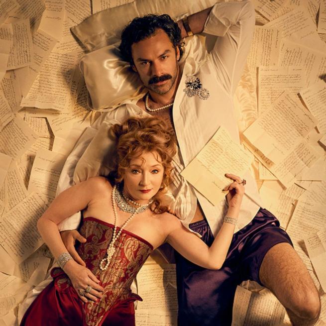 A woman in pearls, diamond jewellery and a red and gold corset lies on a moustached man in an unbuttoned shirt and shorts. Both look up at the camera and lie across sheets of handwritten notes. 