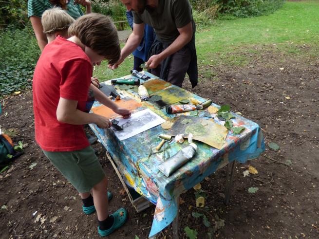 A boy making a monoprint on a table in the woods. There are tubes of ink on the table.