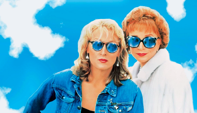 Suzanne Vale played by Meryl Streep and Doris Mann played by Shirley MacLaine. Both have sunglasses on with palm trees. The background is blue sky and clouds. They are both wearing jackets denim and fur coat. 