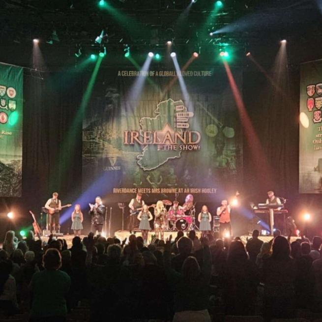 A large band with multiple instrumentalists and singers on stage perform under spotlights to a large crowd.