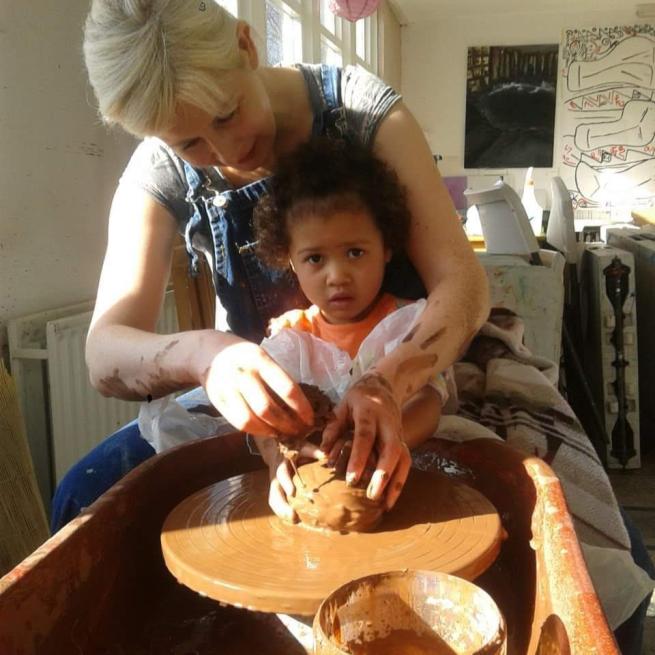 A lady sat at a pottery wheel with a child sat on her lap. They handle some clay.
