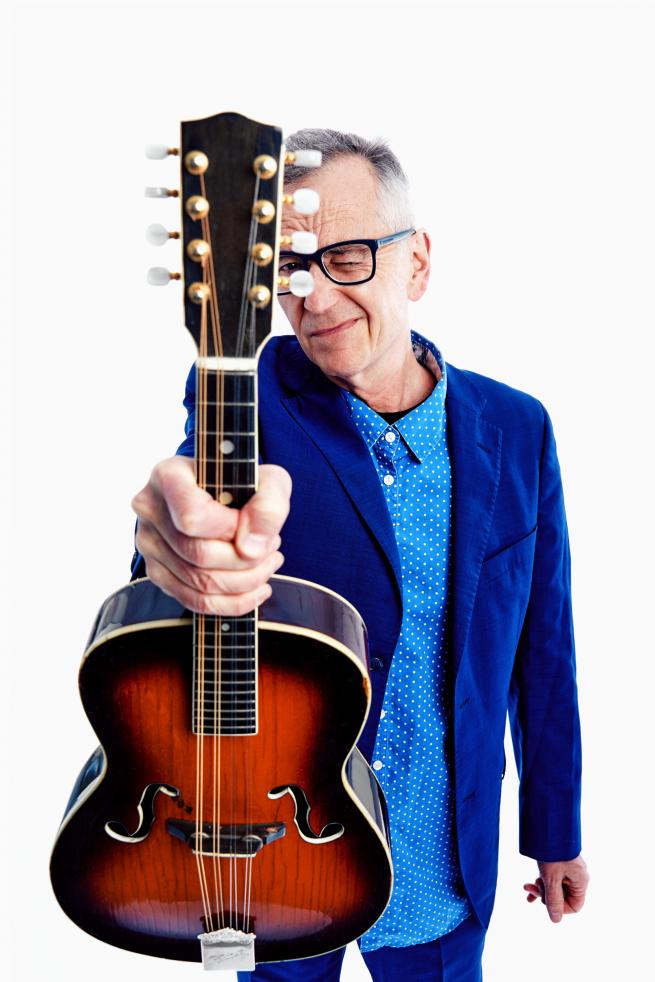 John Hegley is wearing a blue suit and light blue shirt with small polka dots. He is holding up a ukelele towards the camera with one hand whilst peering through the space in its tuning pegs with one eye closed.