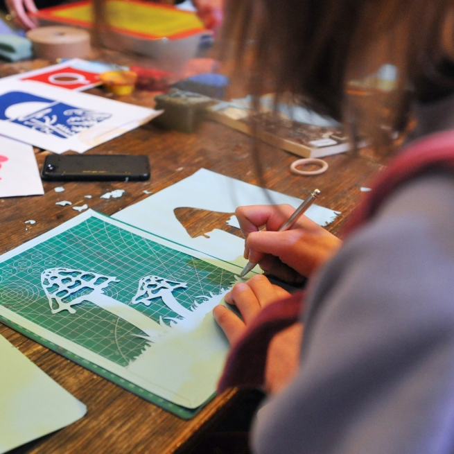 A photograph of a person cutting a paper stencil with a craft knife, the photo is taken over their shoulder