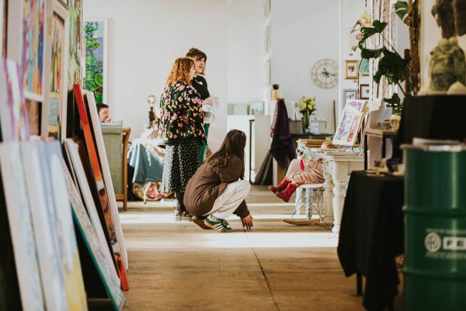 3 studio holders in the Spinning Mills Studios at Sunny Bank Mills chatting at the end of a corridor of studio spaces. The corridor is filled with paintings stacked against walls, plants and ceramic sculptures. 