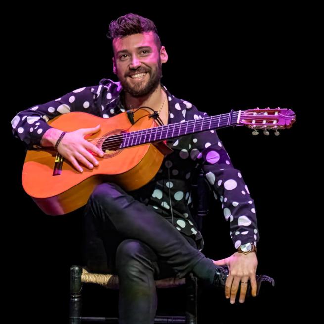 A man smiles on a stool onstage, wearing a polka dot shirt and holding an acoustic guitar.