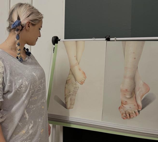 A woman with a grey t-shirt and light hair stands in front of her drawing easel featuring two pencil drawings of ballerina's feet