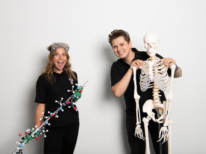 Two people holding scientific objects, one skeleton and one dna stick smiling at the camera.