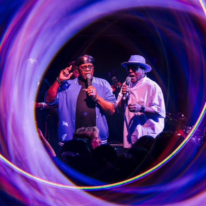 Two singers with microphones perform on stage, surrounded by a soft halo of purple light.