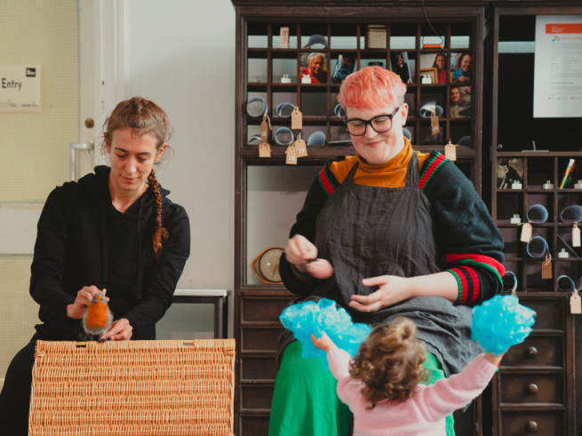 Two female actors, one holding a small robin puppet and the other interacting with a small child holding blue pom poms.