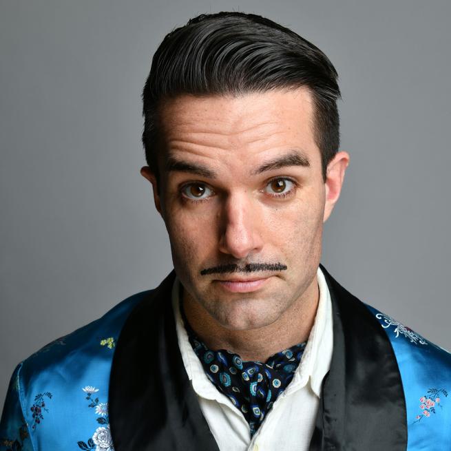 Moustachioed Troy Hawke wears a shiny blue floral smoking jacket and matching neck tie against a plain grey background.