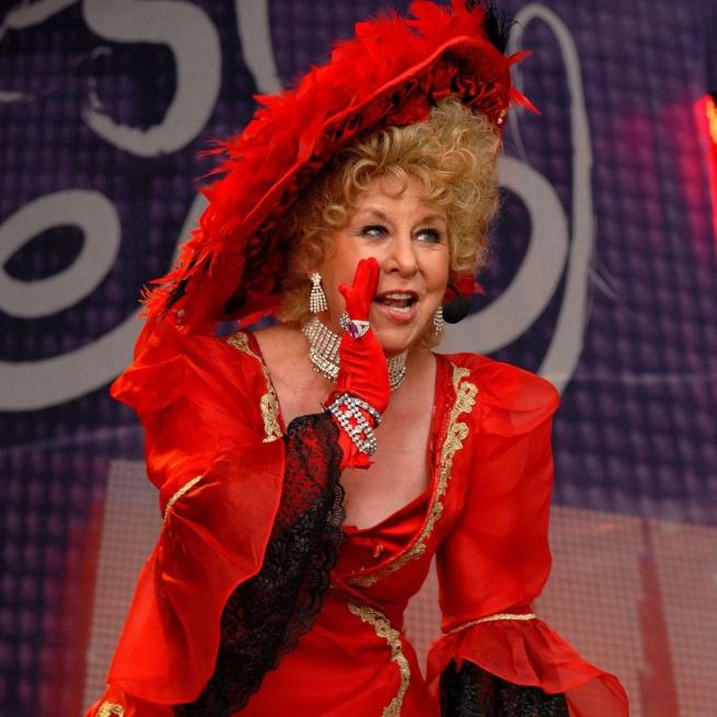 Jan Hunt in The Magnificent Music Hall show, wearing a bright red dress with dangly jewellery, performing on stage.