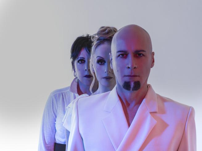 Three people in white clothing stand in a line, facing forward with serious expressions against a light background with blue and pink lighting.