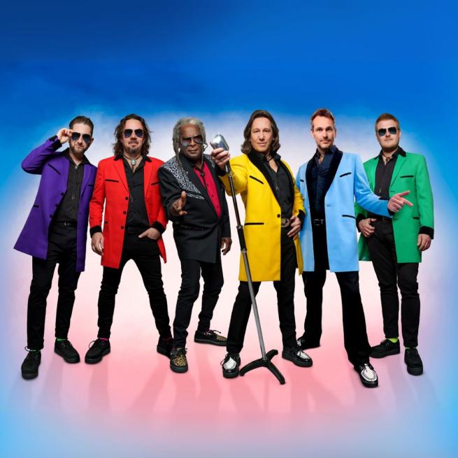 The members of Showaddywaddy stand in brightly coloured suit jackets, posing in sunglasses against a vibrant blur backdrop.