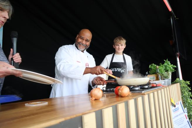 a person in a chefs outfit holding a pan over a grill, with a person in an apron next to them