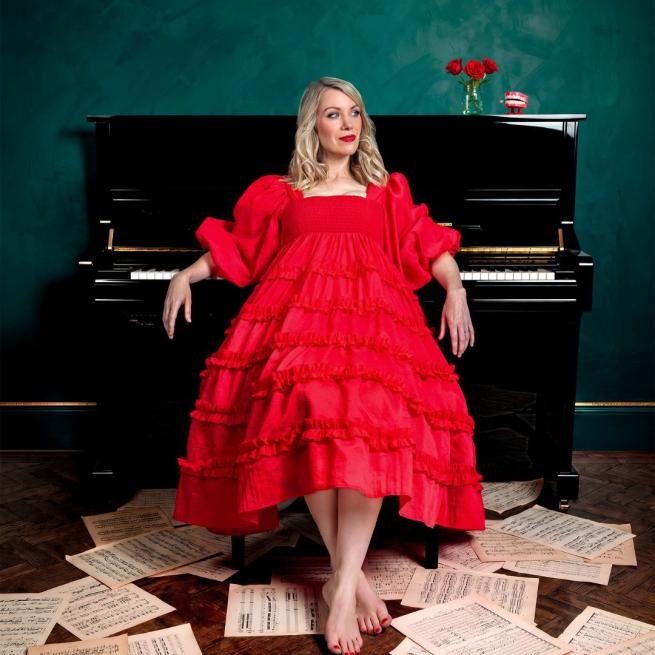 Rachel Parris sat at a grand piano and looking off-camera, wearing a red dress with bare feet on a wooden floor covered with sheet music.