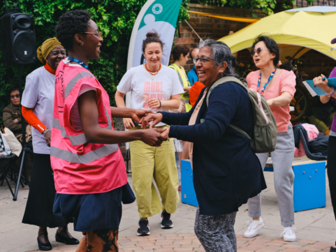 Two women smiling and dancing together at an outdoor community celebration, surrounded by other participants, volunteers and colourful event tents in the Roxby Community Garden.