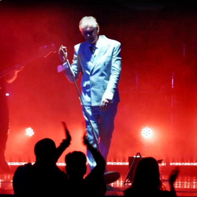 On a hazy stage with red spotlights, a David Bowie tribute act performs in a white suit, accompanied by two guitarists. 