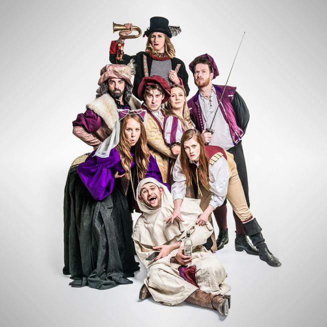 A group of characters in Shakespearean costumes huddle together and pose with quizzical expressions against a blank background.