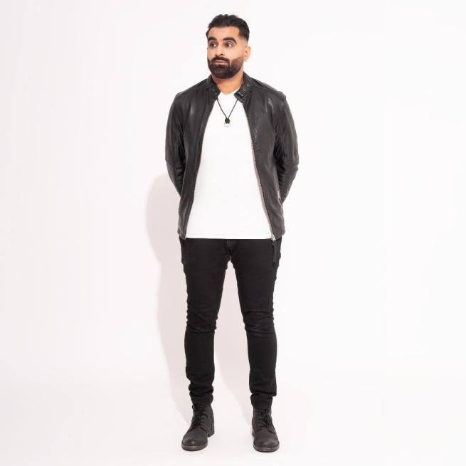 Tez Ilyas stands with his hands behind his back, wearing a leather jacket and white t-shirt as he looks off-camera in front of a white backdrop.