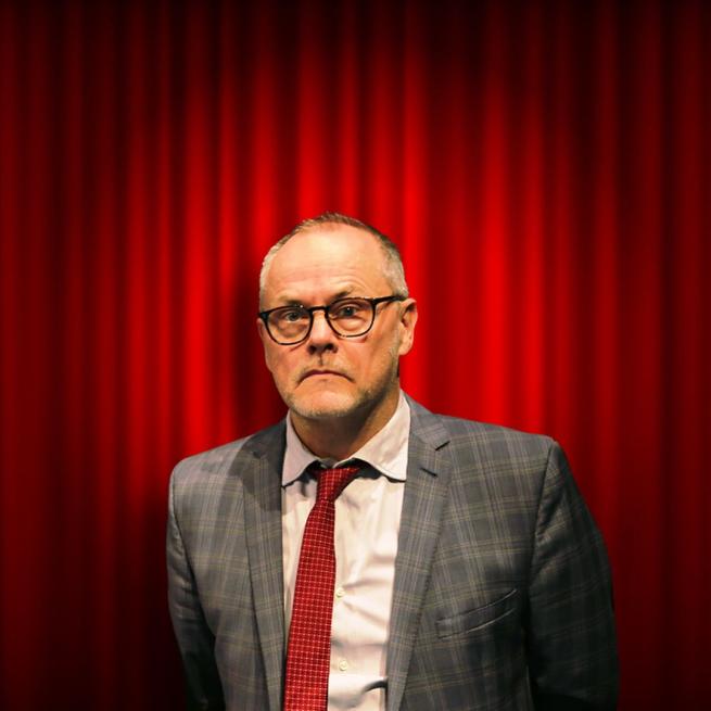 Jack Dee holds a neutral expression, wearing glasses, a grey suit and red tie, positioned in front of a red curtain and lit by a spotlight.