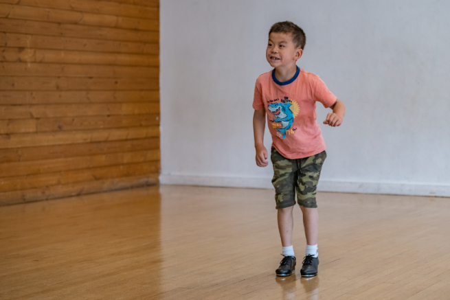 A young, smiley boy is bending his knees wearing tap shoes in a dance studio.